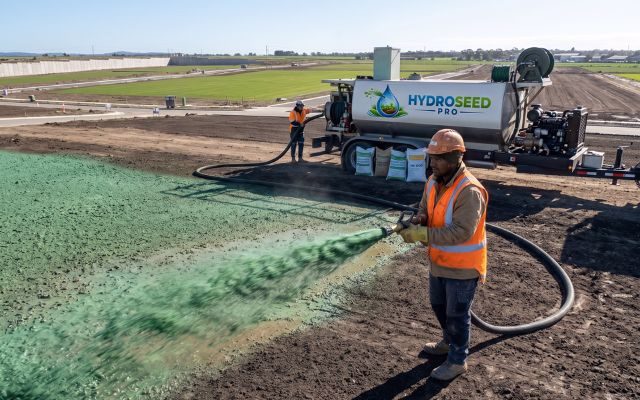 HydroSeed Pro worker spraying green hydroseeding slurry across a large bare dirt site in South East Queensland with branded hydroseeding trailer in background